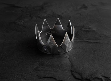 Oxidized silver crown ring with a textured geometric design and studded band, displayed on a dark, cracked slate surface.