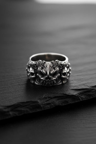 Front view of the silver demon skull ring, showcasing the central demonic face and flanking skulls on a dark slate.