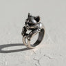 Handcrafted silver cat ring with a playful expression, its paws and tail wrapping the band, on a white textured surface.