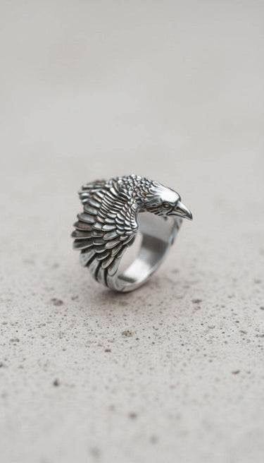 Three-quarter profile of the sterling silver raven ring, highlighting the bird's eye and sharp beak, resting on a stone-like surface.