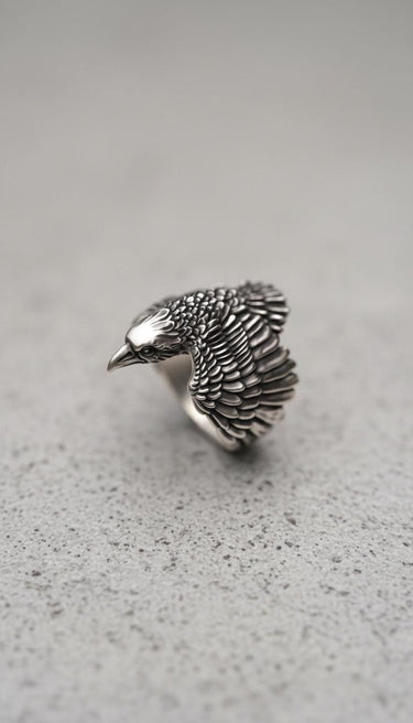 Silver Raven Ring with detailed, oxidized feathers and a polished head, shown at an angle on a textured grey surface.