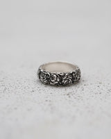Silver rose ring with a detailed, repeating floral pattern shown at an angle on a light gray textured surface.