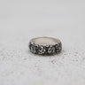Silver rose ring with a detailed, repeating floral pattern shown at an angle on a light gray textured surface.
