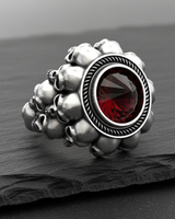 Silver skull ring with a large, round, inverted red stone, shown from the front on a dark, textured slate surface.