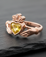 Rose gold Claddagh ring with a vibrant yellow heart-shaped gemstone, resting on a textured dark slate surface for a luxury feel.