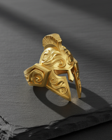 Gold-plated Spartan helmet ring shown from a side angle, highlighting the detailed crest and filigree on a dark background.