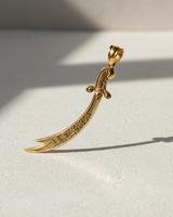Gold vermeil Zulfiqar sword pendant with Arabic calligraphy standing upright on textured surface