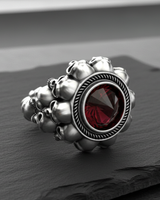 Angled view of the silver skull ring highlighting the clustered skull band and the faceted red gemstone's rope bezel.