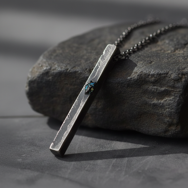 Oxidized silver bar necklace featuring raw texture and iridescent blue gemstone on dark stone background