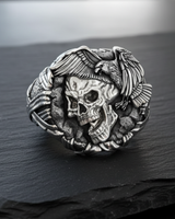 Side angle of sterling silver skull ring showing textured stone band and oxidized raven feather details with brass skull