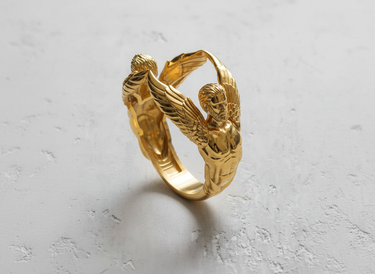 Gold vermeil angel ring sculpture featuring two winged figures on textured white background