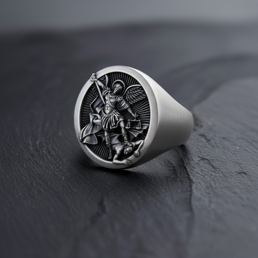 Oxidized sterling silver St Michael Ring featuring the Archangel defeating Satan, shown angled on a dark slate surface.
