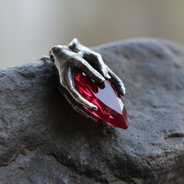 Gothic style 925 sterling silver hands gripping a faceted red garnet drop pendant displayed on natural rock