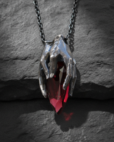 Front view of handcrafted silver skeleton hands holding red gemstone pendant with chain on rock