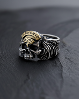 Silver Memento Mori Ring with a gold banner and detailed wings, shown on a dark, textured slate surface from a front angle.