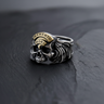 Silver Memento Mori Ring with a gold banner and detailed wings, shown on a dark, textured slate surface from a front angle.