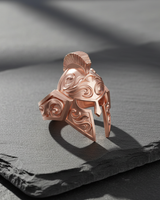 Rose gold plated Silver Spartan Helmet Ring with intricate scrollwork, displayed on a dark slate surface with soft lighting.